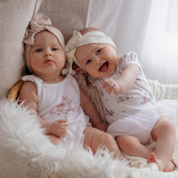 Two babies in white dresses sitting on a fluffy white surface with a neutral background