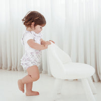 Child in a white romper standing next to a white chair against a white curtain background
