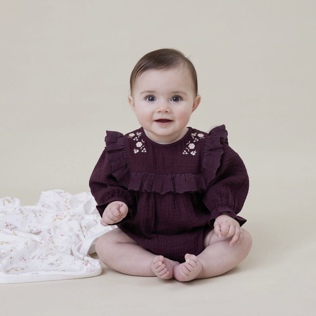 Baby in a dark purple outfit with ruffles sitting on a beige blanket.
