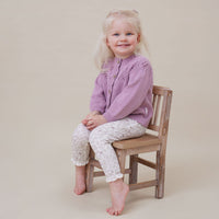 Young girl sitting on a wooden chair wearing a purple sweater and white patterned pants against a beige background.